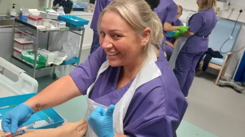 Rachel Ohene-Adjei Rachel wearing purple hospital scrubs and blue surgical gloves. She smiles at the camera and holds a syringe in one hand. 