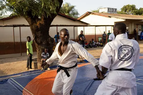 PATRICK MEINHARDT / AFP Two men in white karate suits engage in combat on mats. Spectators look on from the edges.