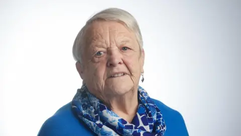 AGE NI a woman against a blank background looking directly at a screen. She is wearing a blue top, a patterned blue and white scarf and drop earrings. Her white hair is styled short and swept over to one side