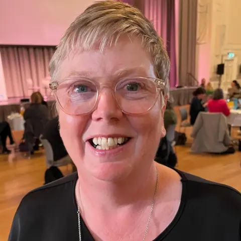 Linda Fay is wearing a black top and large transparent-framed glasses as she smiles at the camera with a hall and a stage behind her. People are sitting at big round tables in the background.