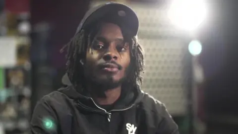 BBC Malachi Saunders, a young man with short dreadlocks wearing a black cap and black hoodie, sitting in front of bright studio lights