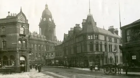Leeds Museum/Leodis A black and white photograph of Leeds around 1900, with people walking on the streets and a horse and cart in the foreground.