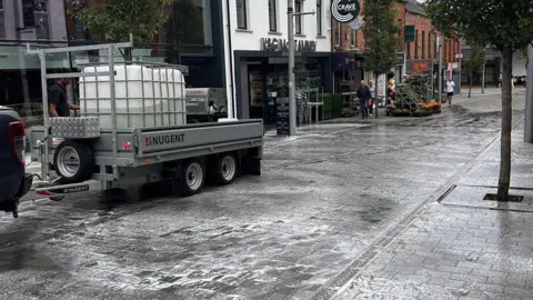A black paved street with businesses down the side of it. There are suds on the ground as people clean up the slurry which was spread.