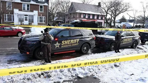 Getty Images Police tape is visible barricading a snow-covered residential street. Two sheriff vehicles are in the foreground with officers positioned in front of them.