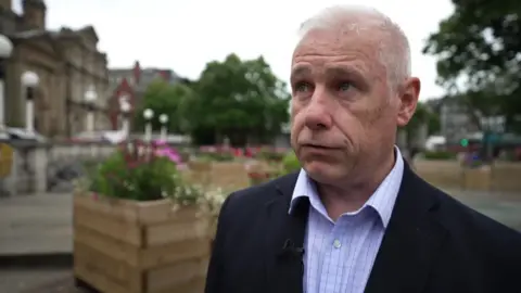 Andy Brown, who has a short white hair and wears a blue suit with a blue shirt, stands in front of the farmers in the square in the center of the town of Soutbort