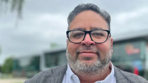 Emma Baugh/BBC A head and shoulders image of Mohammed Jamil. He is wearing a jacket over a shirt and glasses. He is looking directly into the camera.