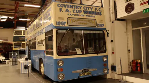 A blue and cream vintage bus, that open top and has writing on it that reads "Coventry City FC, FA Cup Winners. The bus is in a transport museum. 