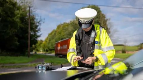 Wiltshire Police  Image shows a police officer looking at their phone and resting again the bonnet of a car. The officer is wearing a police jacket and a bullet-proof vest.