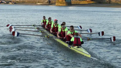 Nine members of Oxford Brookes University Boat Club, all wearing burgundy, row on a stretch of water. Their ores are stripes red, white and blue. Their rowing boat is yellow. Most members are male and are wearing sunglasses.