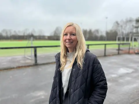 A blonde-haired woman in a dark padded jacket and a grey jumper stands in front of a playing field behind a fence