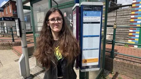 A young woman with long brown hair looks at the camera, with her arms behind her back, standing in front of a bus stop on a high street.
