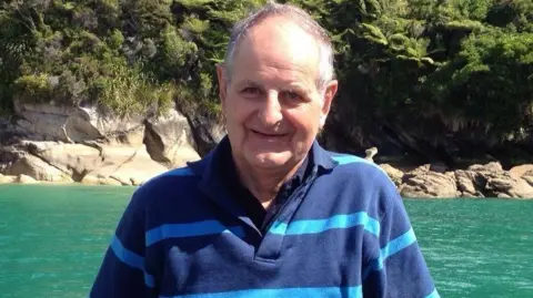 A man with grey hair and a blue striped polo shirt smiling into the camera. He is standing in front of a stretch of turquoise water on a sunny day, with boulders on the water line and a stretch of green trees in the background.