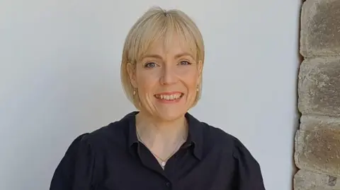 A woman with chin-length blonde hair and a fringe smiles at the camera as she wears a black shirt, silver necklace and makeup.