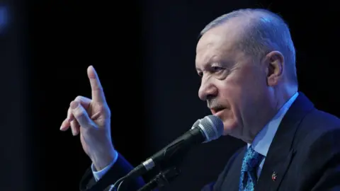 Getty Images Erdogan makes a speech in front of a black background. He is holding his finger in the air and wearing a suit. 