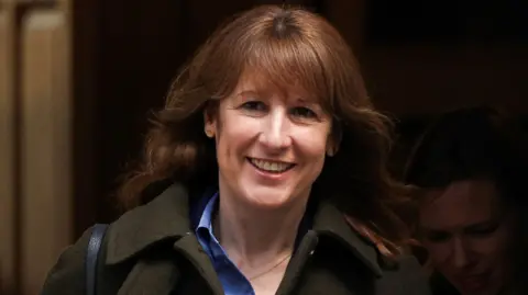 Reuters Chancellor Rachel Reeves smiles as she leaves Downing Street. She wears her light brown hair loose with a fringe. She's wearing a blue shirt and a dark green overcoat.