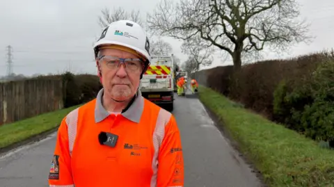 North Northamptonshire Council Chris McGiffin faces the camera wearing a high vis orange clothing and white hard hat. He is on a country lane. Behind him, a crew fixes a pothole on the side of the road.