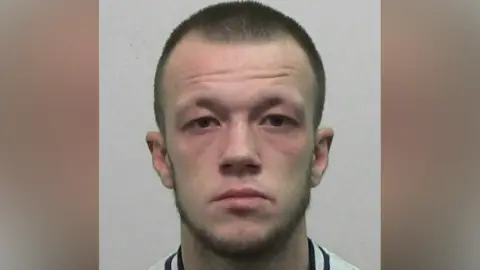 Ryan Scott is looking directly into the camera with a blank look on his face, He has sort dark hair and a wispy beard.