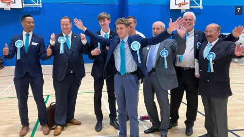 Reform UK candidates in Leicestershire. They all sport blue rosettes and wear suits over white shirts and light blue ties.