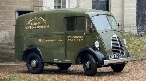 An olive green-coloured original Morris JE van is parked up on gravel in front of a stone building. It has 'Smith & Grieves Builders Painters & Decorators' painted on the side, along with 'Tel: Livingstone 3586' and 'Upper Norwood. London. S E 19'. 