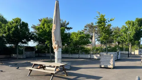 A number of items of street furniture including planters and picnic tables sit in an area close to the River Foyle's edge.