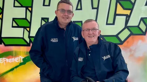 Dave Thompson A father and son pose for a photograph together in front of a graffiti-styled mural. They are both wearing navy jackets branded with their organisation, the Warrington Disability Partnership, in white letters.