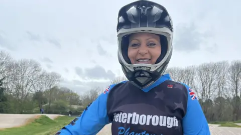Leah Pearson,  in a light and dark blue top saying Peterborough Phantoms. She is wearing a helmet and smiling broadly. Behind her are undulating concerte tracks and leafless trees against a grey sky. 
