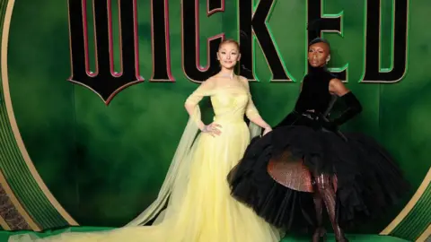 Tim P. Whitby / Stringer/Getty Ariana Grande wearing a yellow dress stands and smiles at the camera next to Cynthia Erivo who wears a puffy black dress. They stand in front of a green backdrop with the words 'Wicked'.