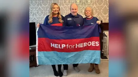 Help for Heroes Anne Duncan, Peter Duncan and Sophie Ombler. They are standing in a living room behind a Help for Heroes navy blue, red and light blue flag which Anne and Sophie are holding. They are each wearing Help for Heroes tops. 