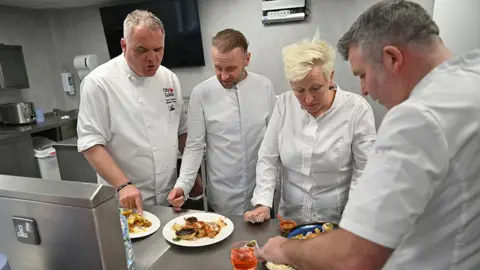 Lisa Godwin-Allen, with short, spiky blonde hair, stands with three male chefs looking plates of food in an industrial steel kitchen at the college. All are wearing chef's whites
