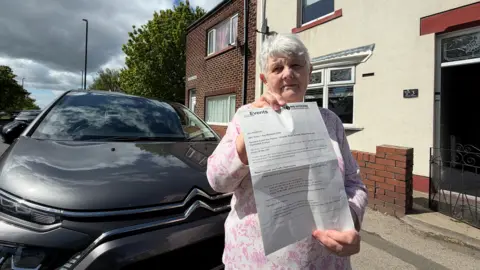 Joan Everett, who is wearing a pink floral dress, stands in front of her and her home while holding up a letter. The letter has been issued by Sunderland Council, which confirms the road will close and she must park at the rear of her home is she is to continue using her car.