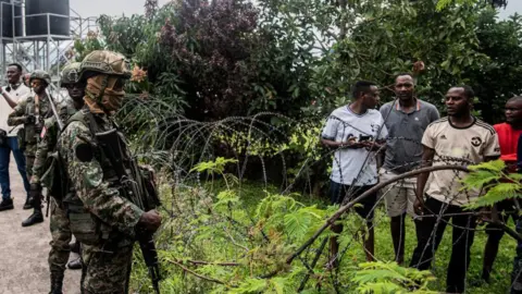 Getty Images Members of the M23 movement, dressed in T-shirts and shorts, stand guard for Rwanda soldiers arriving. Separating them is barbed wire.
