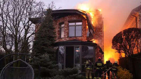 Ukrainian Emergency Service workers stand beneath burning building as fire rages behind them