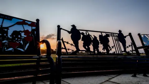 out to grass Silhouettes of soldiers and barbed wire on the back of bench. You can see the sun setting behind and an orange sky. There are small clouds in the sky. One of the three benches has poppy sculptures on the back.