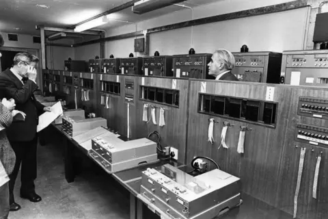 Media Wales Black-and-white photograph of a room filled with telecommunications or computer equipment. Large cabinets line the walls, with control panels, switches and labelled tape or paper strips hanging from them. Several people stand among the equipment, examining documents and the machinery. Long worktables in the foreground hold electronic devices and monitoring equipment, illustrating a mid-20th-century technical operations centre.