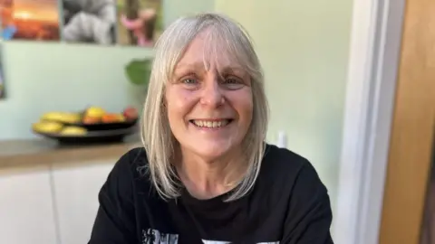 Kate Bradbrook/BBC Woman with below shoulder length blonde hair and black t-shirt smiles at the camera. In the background is a bowl of fruit out of focus