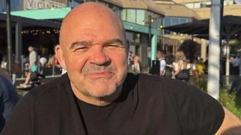 A head and shoulders shot of Paul Hughes. He has greying short stubble, is wearing a dark T-shirt and is about to smile. He is outside and behind him is a shopping centre with people walking around. 