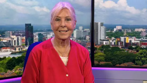 Lorraine Pugh smiling at the camera - she has pale blond hair tied up and wears a bright fuschia cardiagan.