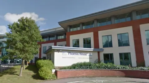 Phoenix House district council offices in Tiverton. A council building made of white and red brick. It has sets of four windows positions in a square and a small outbuilding with the logo written on it. There is a tree to the left of the image. 