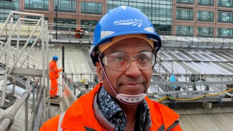 A man is in a blue hard hat and an orange hi viz jacket standing in front of a roof