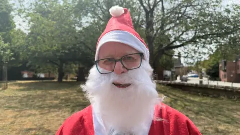 A man looking at the camera. He is wearing a Father Christmas outfit – a red and white hat and jacket. He has glasses and a fake white beard. There is grass and trees in the background.