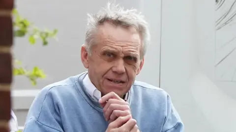 An elderly man wearing a blue jumper over a white suit shirt. He has white hair and is standing in front of a white background with red bricks in the foreground to the left.