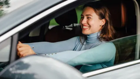 a young woman behind the wheel of a car, grinning broadly