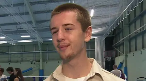 A man with a short beard on his chin, wearing a white collared shirt, smiles in the direction of the camera. He appears to be stood in a gym or hall.