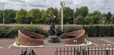 Sunderland AFC Heaven Branch A memorial area in a car park with two curved brick walls and a statue of in the middle. There are bushes, grass and trees. There is also a dark fence.