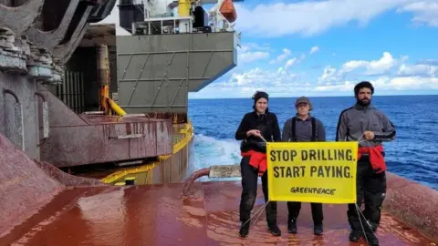 Greenpeace Three Greenpeace activists in dark trousers and fleeces standing on a large ship which is on the sea. They are holding a yellow banner that reads Stop Drilling Start Paying - Greenpeace. 