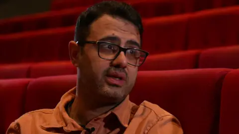 Arash Sedighi, now in his 40s, with dark brown hair, light stubble, black framed glasses and an orange overshirt, sitting in a red cinema seat looking past the camera at a screen. 