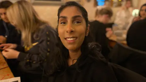 Kaveeta Sehjal is wearing a black jumper and has black long hair. Women can be seen sitting at tables either side of her