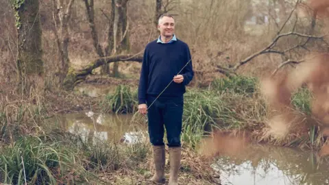 Ben Goldsmith Ben Golding stands in a wild-looking forested area. The ground is overgrown and there is some water on the ground. He is wearing wellies on a patch of dry ground.