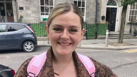 Orlaith Conner tiene el pelo rubio/castaño recogido y con la raya en medio. Está sonriendo y de pie frente a un edificio.