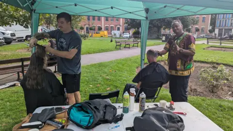 Handout Two men cutting hair outside under a marquee